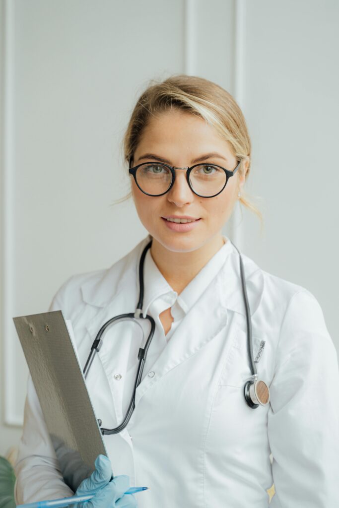 Smiling female healthcare professional in white coat holding clipboard indoors.