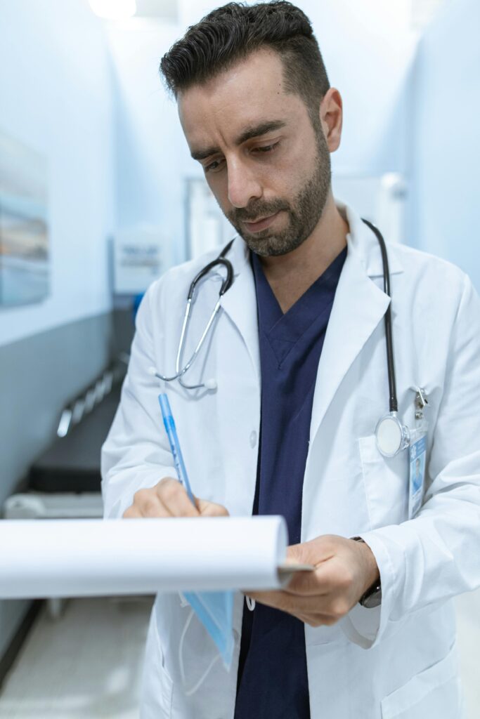 A male doctor writing in a medical chart in a hospital hallway.