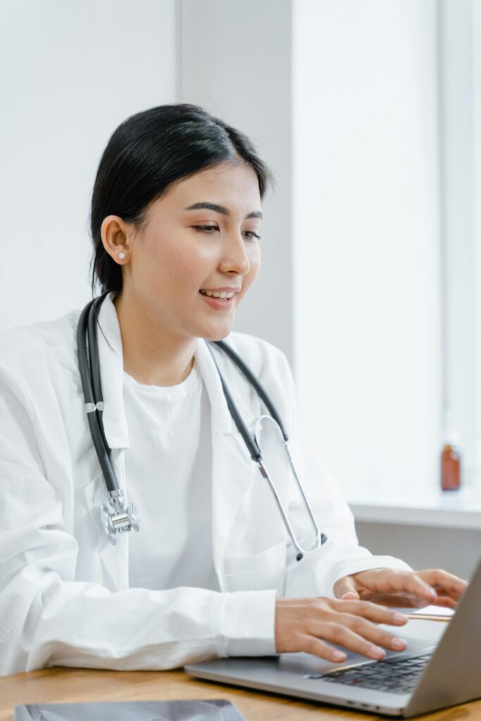 Asian female doctor in a white coat typing on a laptop during a telemedicine session.